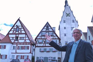 Eine Person mit Brille, im Sakko und blauem Hemd steht vor Fachwerkhäusern und einem Turm mit Uhr und zeigt darauf. Vor den Häusern stehen auf einer gepflasterten Fläche vier Autos geparkt.