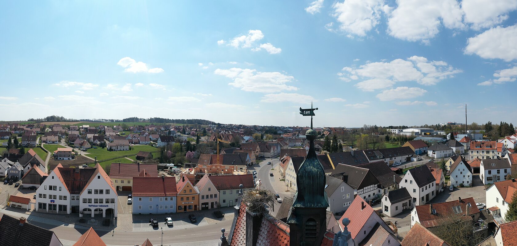 Panoramafoto Monheim mit Storchennest auf dem Oberen Stadttor Panoramablick auf eine Kleinstadt mit roten Dächern, umgeben von Feldern und Wäldern unter blauem Himmel mit Wolken.