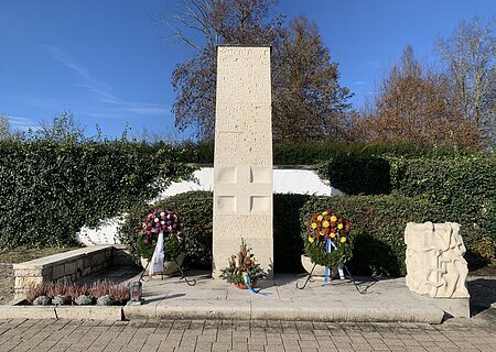 Steinmonument mit zwei Blumengestecken und einer Skulptur, umgeben von Hecken und Bäumen. Friedhof Monheim Volkstrauertag 2024