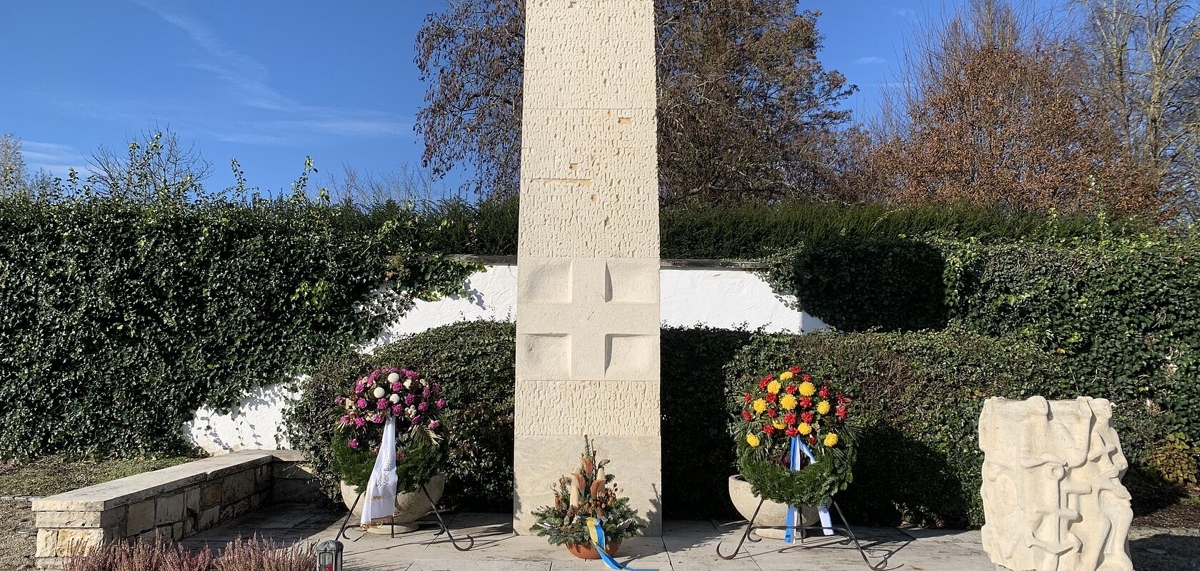 Steinmonument mit zwei Blumengestecken und einer Skulptur, umgeben von Hecken und Bäumen. Friedhof Monheim Volkstrauertag 2024
