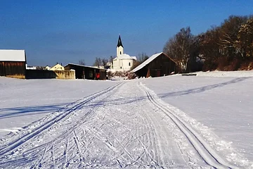 Verschneite Landschaft mit Skispuren, ein Ort zum Langlaufen in der Monheimer Alb. Im Hintergrund Gebäude, eine Kirche und blauer Himmel zu sehen.