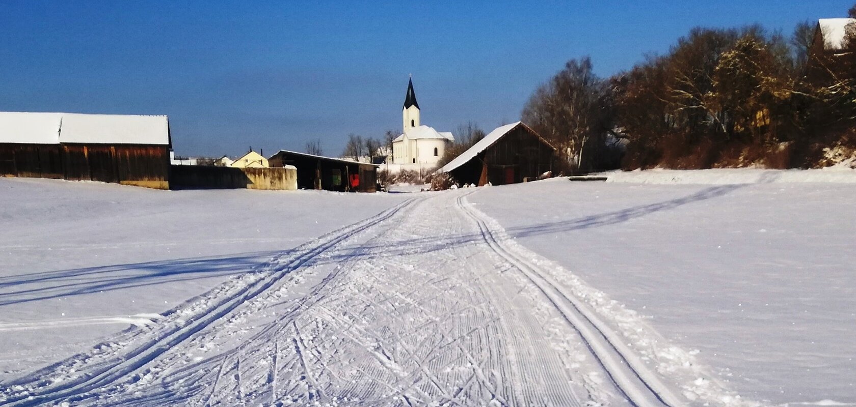 Verschneite Landschaft mit Skispuren, ein Ort zum Langlaufen in der Monheimer Alb. Im Hintergrund Gebäude, eine Kirche und blauer Himmel zu sehen.