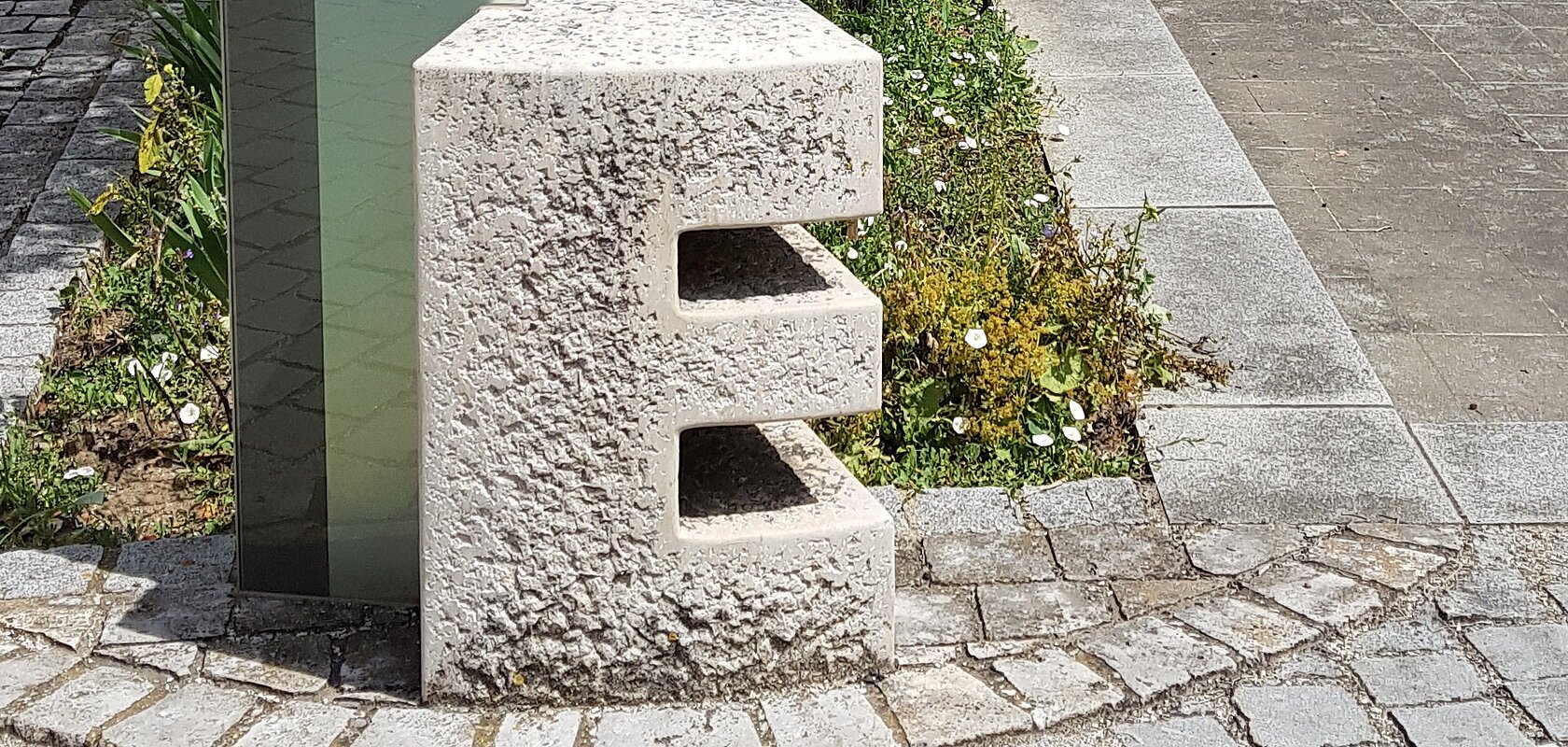 Steinskulptur des Buchstabens "E", dahinter ein Informationsschild und ein Baum mit einem dahinter liegendem Beet. Umgeben von einem gepflastertem Platz, mit einem historischen Gebäude im Hintergrund.