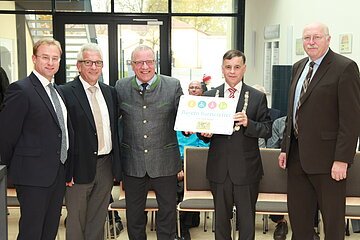 Fünf Männer in Anzügen stehen in einem Raum, einer hält ein Schild mit der Aufschrift "Bayern barrierefrei". Im Hintergrund große Eingangstür aus Glas.