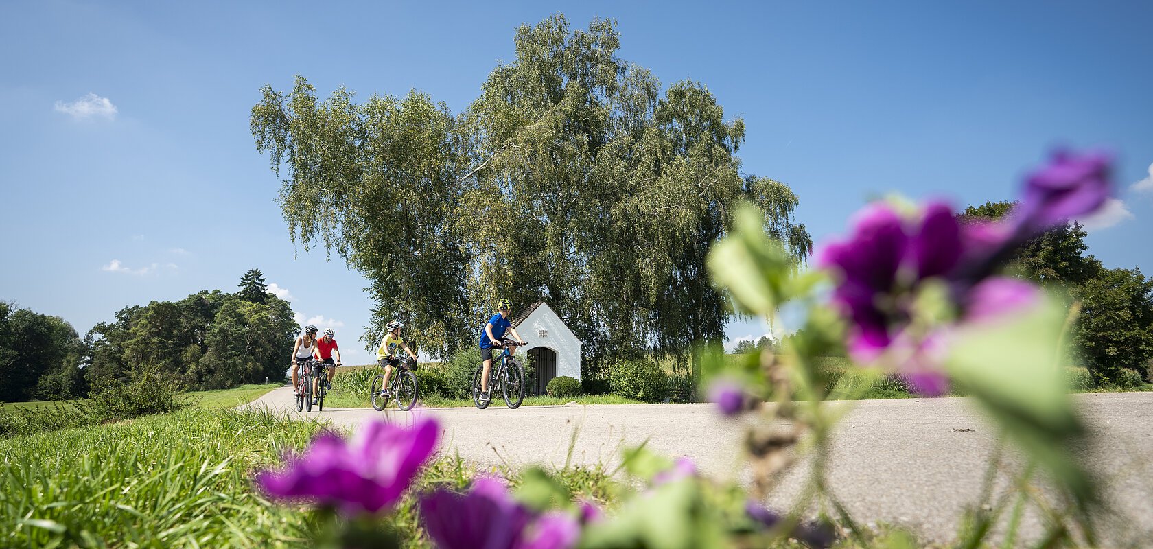 Kapelle Warching, Monheim Drei Personen fahren auf Fahrrädern auf einem Weg, im Hintergrund Bäume und eine kleine Kapelle. Im Vordergrund unscharfe Blumen.