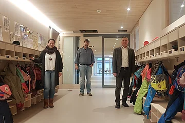 Über den neuen Kindergarten freuen sich die Leiterin Susanne Utjesinovic, Stadtbaumeister Richard Meyer und Bürgermeister Günther Pfefferer (v.l.n.r) Drei Personen stehen in einem Flur mit Kindergarderoben und bunten Jacken. Im Hintergrund ist ein Raum mit Tischen und kleinen Stühlen sichtbar.