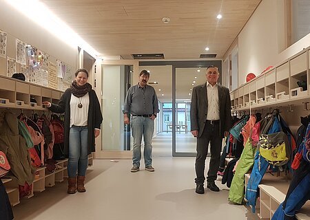 Drei Personen stehen in einem Flur mit Kindergarderoben und bunten Jacken. Im Hintergrund ist ein Raum mit Tischen und kleinen Stühlen sichtbar.