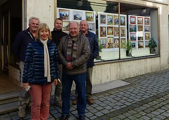 Kunstmaler Hans Mayer im Schaufenster mit Helfer vom Hama Messebau Fünf Personen stehen vor einem Schaufenster mit vielen Gemälden. Pflastersteinstraße im Vordergrund.
