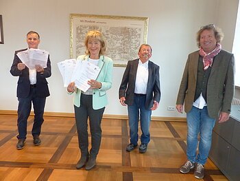 Ziehung der Gewinner zum Monheimer Schaufensterbummel mit Kunst-Rallye Vier Personen stehen in einem Raum mit einem Holzboden, zwei halten Dokumente in den Händen. Im Hintergrund hängt eine Karte von Alt-Monheim im goldenen Rahmen eingerahmt.