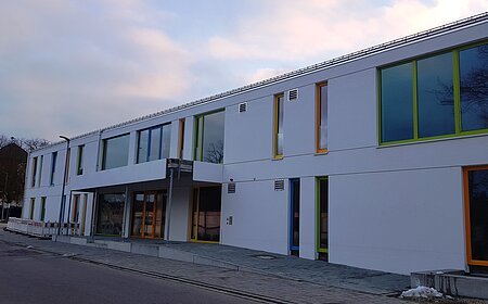 Modernes zweistöckiges Gebäude, der städtische Kindergarten Monheim in weiß, mit vielen bunten Fensterrahmen an einer Straße bei bewölktem Himmel.