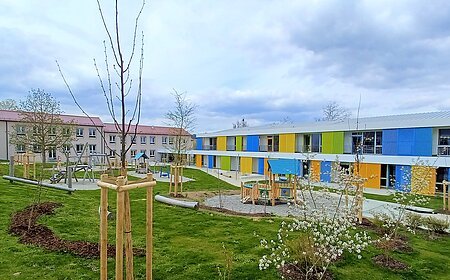 Zwei Gebäude des städtischen Kindergartens Monheim, eins davon mit einer bunten Fassade, davor ein Garten mit Spielgeräten Bäumen und Rasenfläche, bewölkter Himmel.
