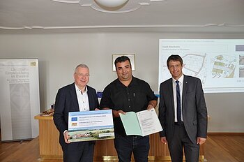 ELER Förderbescheid erhalten für das Projekt "Dorferneuerung - Straßenausbau Flotzheim" Drei Männer stehen in einem Raum, einer hält ein Plakat, ein anderer ein Dokument in den Händen. Im Hintergrund ein Banner vom Amt für Ländliche Entwicklung in Bayern und eine Präsentation an der Wand mit einer Karte.