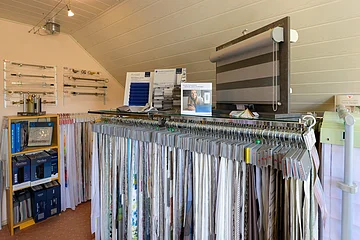 Ein Raum mit Stoffmustern von Gardinen auf Kleiderbügeln, Rollos und Vorhangstangen an der Wand. Verschiedene Displays zeigen Produkte.