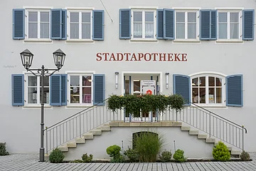 Stadtapotheke - Eingang Gebäude mit blauen Fensterläden, rotem Schriftzug "STADTAPOTHEKE", einer Treppe und einer Laterne mit einem bepflanzten Beet davor.