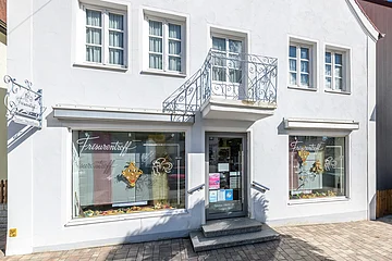 Fassade eines Friseursalons mit Fenstern, zwei Schaufenstern und einem Eingang. Am grau-blauem Gebäude die Aufschrift "Frisurentreff" und oberhalb der Eingangstür ein kleiner Balkon. Vor dem Gebäude ein gepflasterter Fußgängerweg.