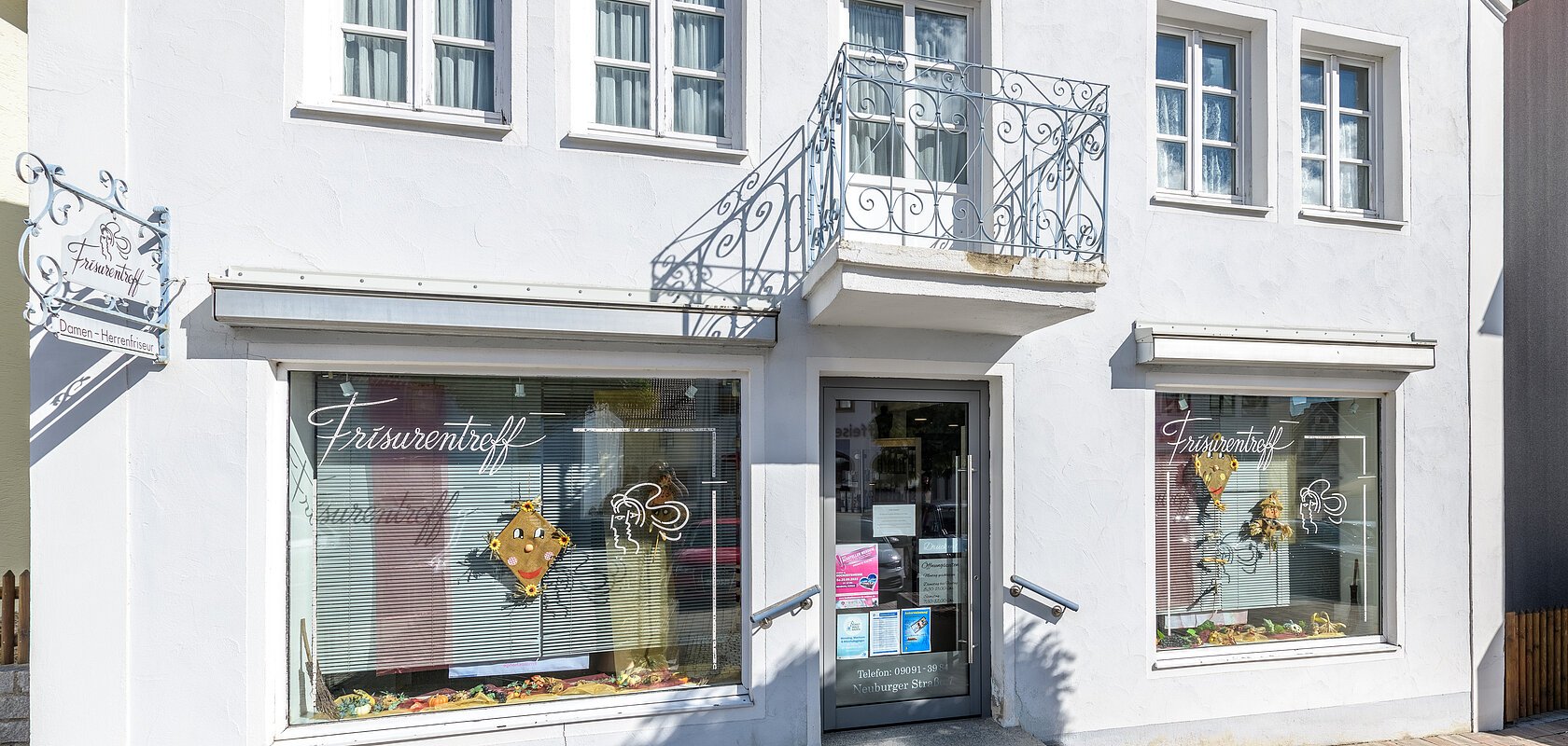 Fassade eines Friseursalons mit Fenstern, zwei Schaufenstern und einem Eingang. Am grau-blauem Gebäude die Aufschrift "Frisurentreff" und oberhalb der Eingangstür ein kleiner Balkon. Vor dem Gebäude ein gepflasterter Fußgängerweg.