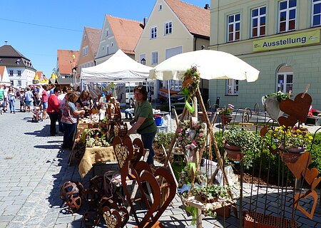 Kunsthandwerker präsentieren sich beim KunstHandWerkMarkt Monheim Menschen besuchen einen Markt mit Ständen, die Kunsthandwerk und Pflanzen auf einem gepflasterten Platz verkaufen. Grünes Gebäude mit einem Banner "Ausstellung" darauf im Hintergrund und blauer Himmel.