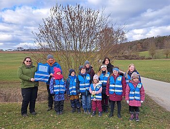 Monheim summt! - ProGeMo spendet Warnwesten Eine Gruppe von Erwachsenen und Kindern in blauen Westen steht im Freien auf einem Feld vor einem Baum. Zwei erwachsen Personen halten zusammen eine blaue Weste in den Händen. Rechts von ihnen verläuft ein Schotterweg. Im Hintergrund blauer Himmel.