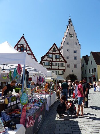 KunstHandWerkMarkt Monheim Markttreiben mit Marktständen und Menschen auf einem gepflasterten Altstadt-Platz vor Fachwerkhäusern und einem Turm mit einer Uhr, bei klarem Himmel.