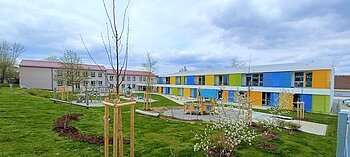 Städtische Kindertagesstätte Monheim Zwei Gebäude des städtischen Kindergartens Monheim, eins davon mit einer bunten Fassade, davor ein Garten mit Spielgeräten Bäumen und Rasenfläche, bewölkter Himmel.