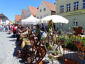 Menschen besuchen einen Markt mit Ständen, die Kunsthandwerk und Pflanzen auf einem gepflasterten Platz verkaufen. Grünes Gebäude mit einem Banner "Ausstellung" darauf im Hintergrund und blauer Himmel.