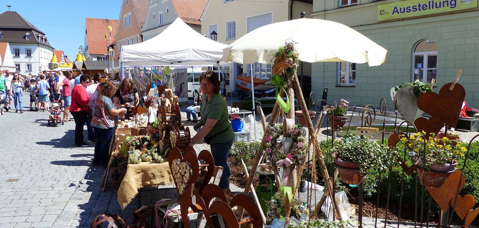 Menschen besuchen einen Markt mit Ständen, die Kunsthandwerk und Pflanzen auf einem gepflasterten Platz verkaufen. Grünes Gebäude mit einem Banner "Ausstellung" darauf im Hintergrund und blauer Himmel.