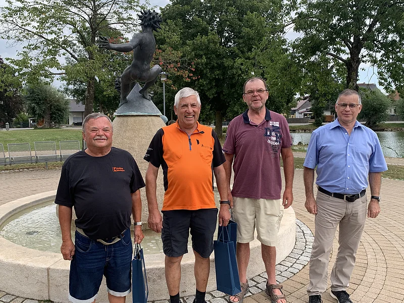 Vier Männer stehen vor einem Brunnen mit einer bronzenen Skulptur auf einem gepflastertem Platz. Im Hintergrund sind Bäume und ein kleiner Weiher.