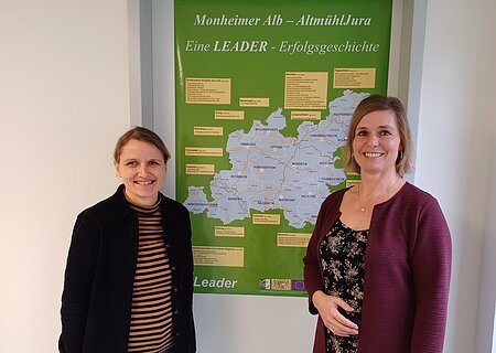 Zwei lächelnde Personen stehen vor einem grünen Plakat mit einer Karte der Region Monheimer Alb-AltmühlJura. Umgeben von einer weißen Wand.