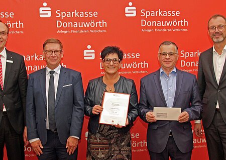 Fünf Personen stehen vor einem roten Hintergrund mit dem Logo der Sparkasse Donauwörth. Eine Person hält ein Zertifikat und eine weitere Person einen Brief in den Händen.