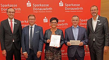 Monheim summt - Nachhaltigkeitspreis 2023 Fünf Personen stehen vor einem roten Hintergrund mit dem Logo der Sparkasse Donauwörth. Eine Person hält ein Zertifikat und eine weitere Person einen Brief in den Händen.