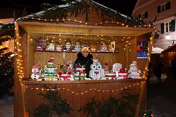 Holzstand auf einem Weihnachtsmarkt mit Windeltorten. Dekoriert mit Tannenzweigen und Lichterketten, eine Person in einer schwarzen Jacke steht dahinter.