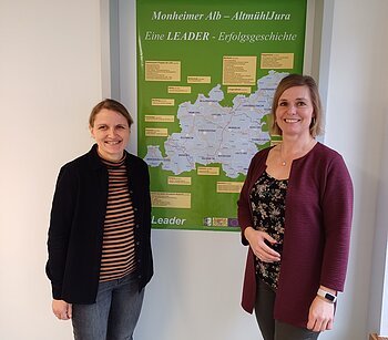 LAG Geschäftsstelle Monheim Zwei lächelnde Personen stehen vor einem grünen Plakat mit einer Karte der Region Monheimer Alb-AltmühlJura. Umgeben von einer weißen Wand.
