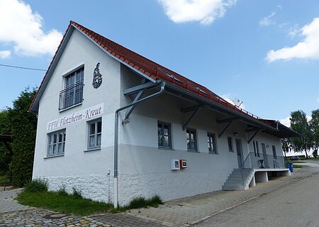 Weißes Gebäude mit rotem Dach, einer Treppe und einem Defibrillator. steht auf einem gepflasterten Grundstück. Auf dem Gebäude der Schriftzug "FFW Flotzheim-Kreut" an der Fassade. Blauer Himmel und Bäume im Hintergrund.