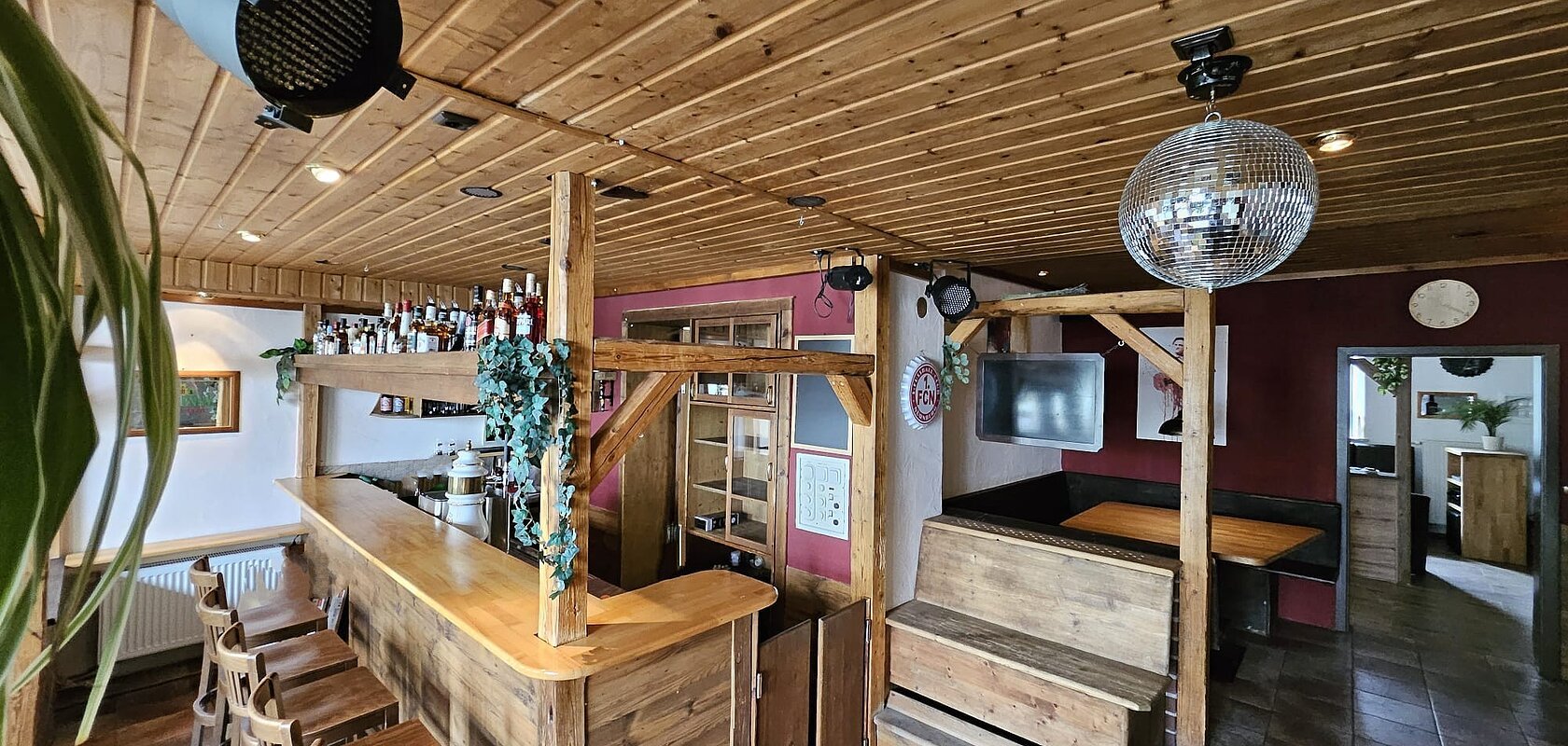 Monheimer Nest - Theke Ein rustikaler Barbereich mit Holzmöbeln, einer Theke, Barhockern, einem schwarzen Scheinwerfer und einer Discokugel an der Decke.