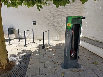 Fahrradservice-Station mit Werkzeugen und Fahrradständern auf einem gepflasterten Gehweg unter einem Baum. Umgeben von einer weißen Mauer.
