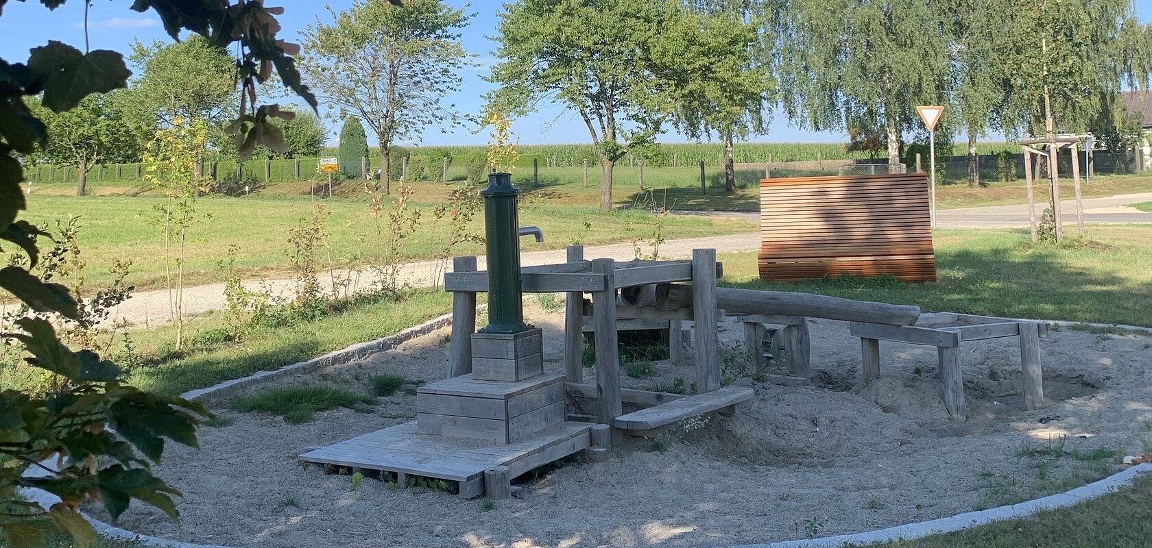 Rund gestalteter Wasserspielplatz mit Holz-Wasserpumpe und Sandbereich, umgeben von einer Rasenfläche. Im Hintergrund Bäume und blauer Himmel.
