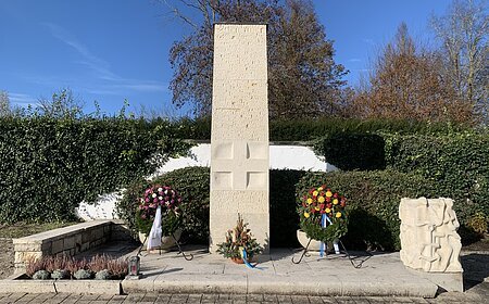 Volkstrauertag 2024 - Friedhof Monheim Steinmonument mit zwei Blumengestecken und einer Skulptur, umgeben von Hecken und Bäumen. Friedhof Monheim Volkstrauertag 2024