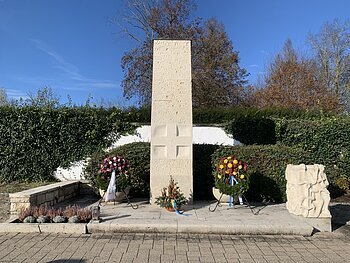 Volkstrauertag 2024 - Friedhof Monheim Steinmonument mit zwei Blumengestecken und einer Skulptur, umgeben von Hecken und Bäumen. Friedhof Monheim Volkstrauertag 2024