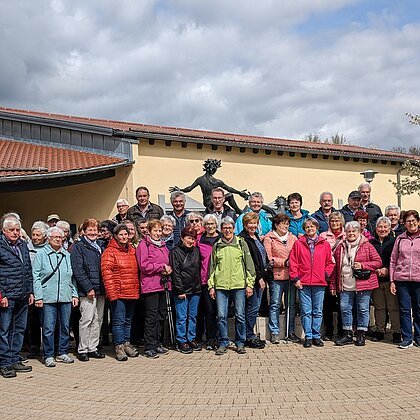 Große Gruppe von Menschen steht vor einem gelben Gebäude mit rotem Dach und einer Statue hinter Ihnen. Im Hintergrund ein Baum auf einer Wiese und bewölkter Himmel.