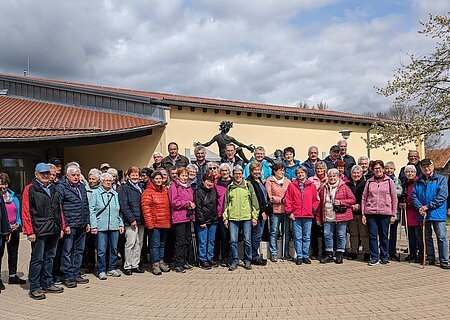 Große Gruppe von Menschen steht vor einem gelben Gebäude mit rotem Dach und einer Statue hinter Ihnen. Im Hintergrund ein Baum auf einer Wiese und bewölkter Himmel.