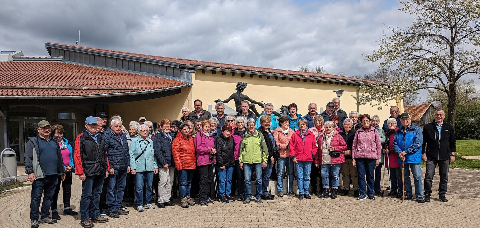Große Gruppe von Menschen steht vor einem gelben Gebäude mit rotem Dach und einer Statue hinter Ihnen. Im Hintergrund ein Baum auf einer Wiese und bewölkter Himmel.