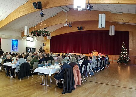 Seniorenweihnachtsfeier Stadt Monheim & Bayer. Rotes Kreuz 2017 Menschen sitzen in mehreren Reihen an weißgedeckten Tischen in einem Saal mit einer Holzbalkendecke und Weihnachtsdekoration. Eine Bühne mit einem roten Vorhang ist im Hintergrund.