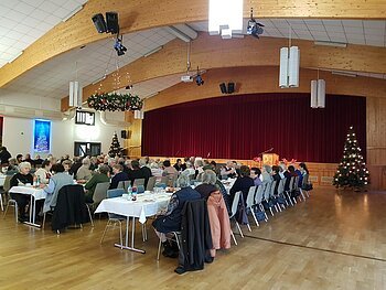 Menschen sitzen in mehreren Reihen an weißgedeckten Tischen in einem Saal mit einer Holzbalkendecke und Weihnachtsdekoration. Eine Bühne mit einem roten Vorhang ist im Hintergrund.