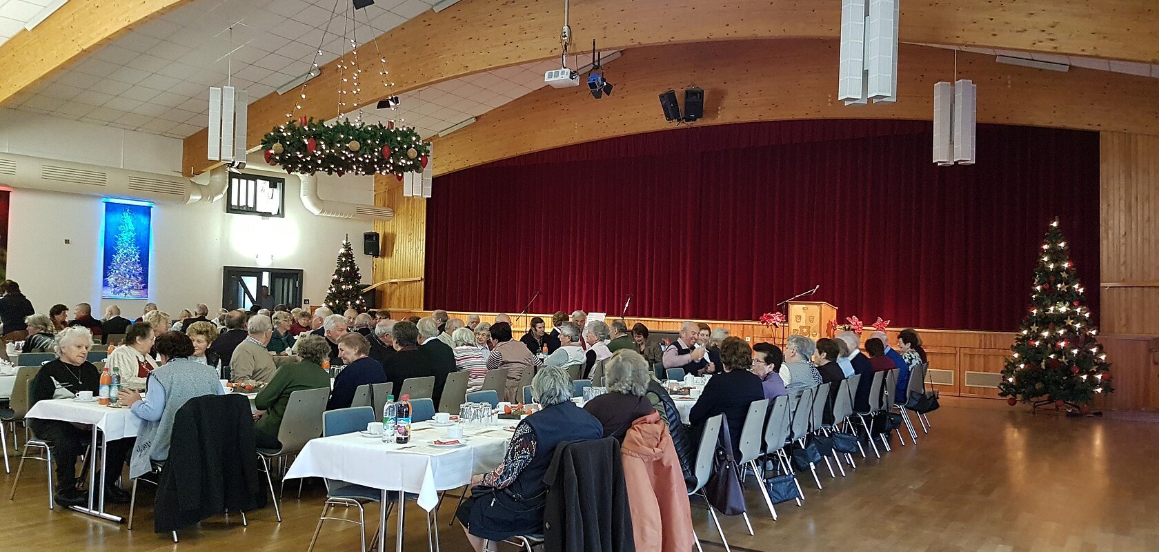 Menschen sitzen in mehreren Reihen an weißgedeckten Tischen in einem Saal mit einer Holzbalkendecke und Weihnachtsdekoration. Eine Bühne mit einem roten Vorhang ist im Hintergrund.