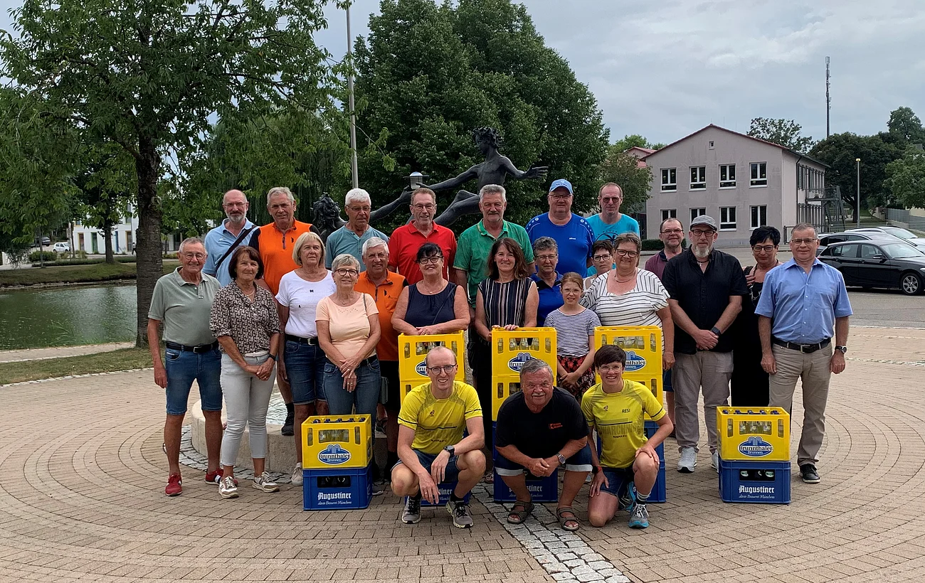 Gruppe von Personen steht draußen auf einem gepflasterten Platz. Vor ihnen sind Getränkekisten in blau und gelb aufgestellt. Im Hintergrund sind Bäume, ein Weiher, ein Gebäude und ein Auto.