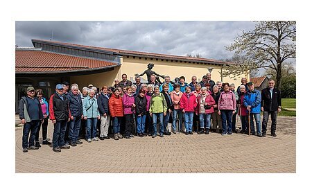 Große Gruppe von Menschen steht vor einem gelben Gebäude mit rotem Dach und einer Statue hinter Ihnen. Im Hintergrund ein Baum auf einer Wiese und bewölkter Himmel.