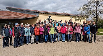 Große Gruppe von Menschen steht vor einem gelben Gebäude mit rotem Dach und einer Statue hinter Ihnen. Im Hintergrund ein Baum auf einer Wiese und bewölkter Himmel.