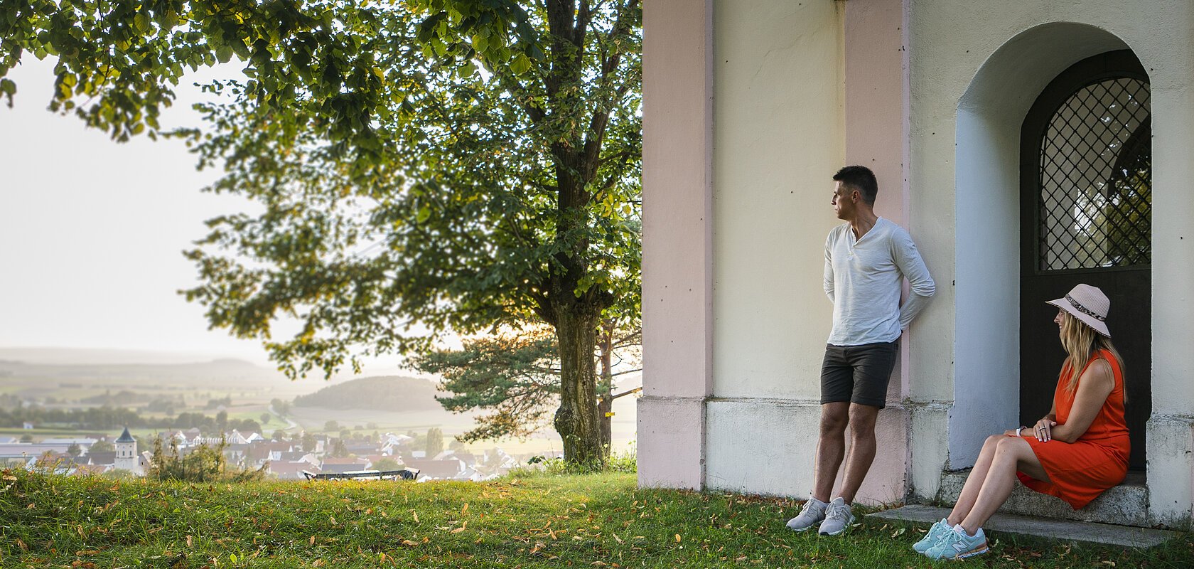 Mann lehnt an Hauswand, Frau sitzt in Türbogen, große Bäume, Dorf im Hintergrund bei Tageslicht.