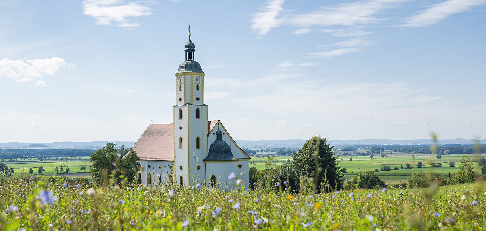 Kirche mit Turm hinter blühendem Feld unter blauem Himmel mit Wolken in ländlicher Umgebung