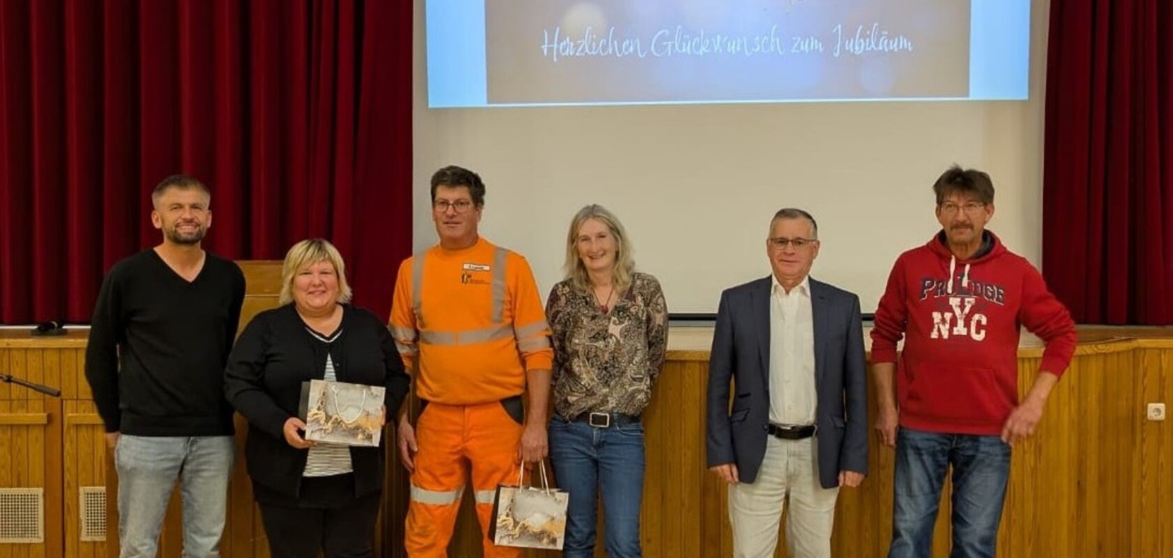 Sechs Personen stehen nebeneinander auf einer Bühne vor rotem Vorhang und Leinwand mit Jubelbild und Text. Es handelt sich um den Personalrat der Stadt Monheim und geehrte Jubilare der Stadt Monheim